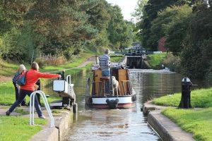 On the Inland Waterways. A narrowboat being steered by a man wearing jeans and a striped polo top as it leaves a lock heading up the Audlem lock flight. A yellow labrador beside him. A lady in an orange top and jeans is starting to close the gate, whilst another lady in a striped top and dark trousers is winding down the paddle gear.