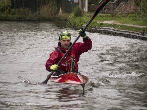 A bearded man in a yellow helmet and red waterproofs powering a red and white kayak