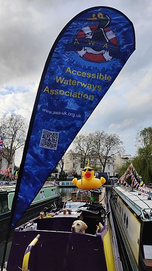 Moored up in the pool, Little Venice, at Canalway Cavalcade. A big "Tear Drop" flag with the Accessible Waterways Association logo at the top towers over the boat. A huge inflatable rubber duck is on the roof!