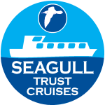 Seagull Trust logo. A circle, the top half of which is light blue, the bottom dark blue joined in a slightly wavy line to represent water. At the top is a drawing of a seagull in a dark blue circle. On the join is a drawing of a white boat. The white words "Seagull Trust Cruises" is in the bottom half.