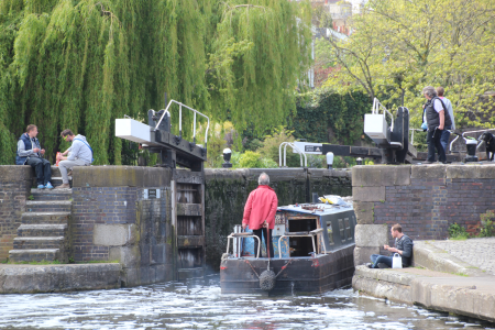 A boat entering a lock to go up. There is a person sat on the lower lock landing, and a couple on the top of the lock steps opposite enjoying a picnic. Two people are on the towpath side ready to close the lock gates and work the lock.