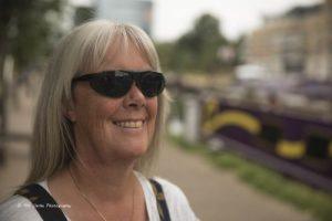 Picture of Tracey Clarke, with long silver hair and dark glasses. The background is out of focus, but a canal, with a purple narrowboat with a wavy yellow band can be seen.