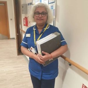 Rekha Vijayshankar - a Research Nurse for Marie Curie. She is wearing a blue nurses uniform with elbow length sleeves and holding a full lever-arch folder. She has shoulder length curly grey hair and glasses.