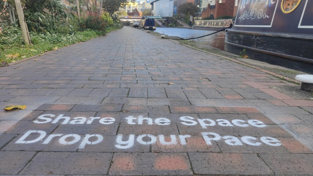 White stencilled sign "Share the Space Drop your Pace" on a brick towpath.