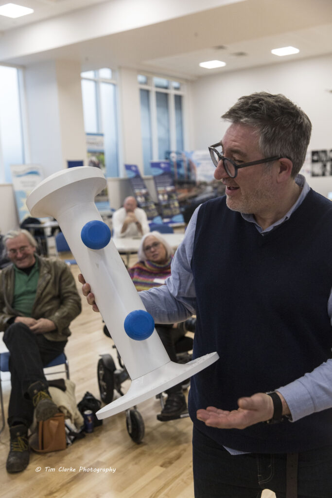 Dick Vincent showing off the new prototype accessible mooring bollard. It is approx 2' tall, white, with a circular base and domed top. There are two arms at different heights to hold mooring ropes.