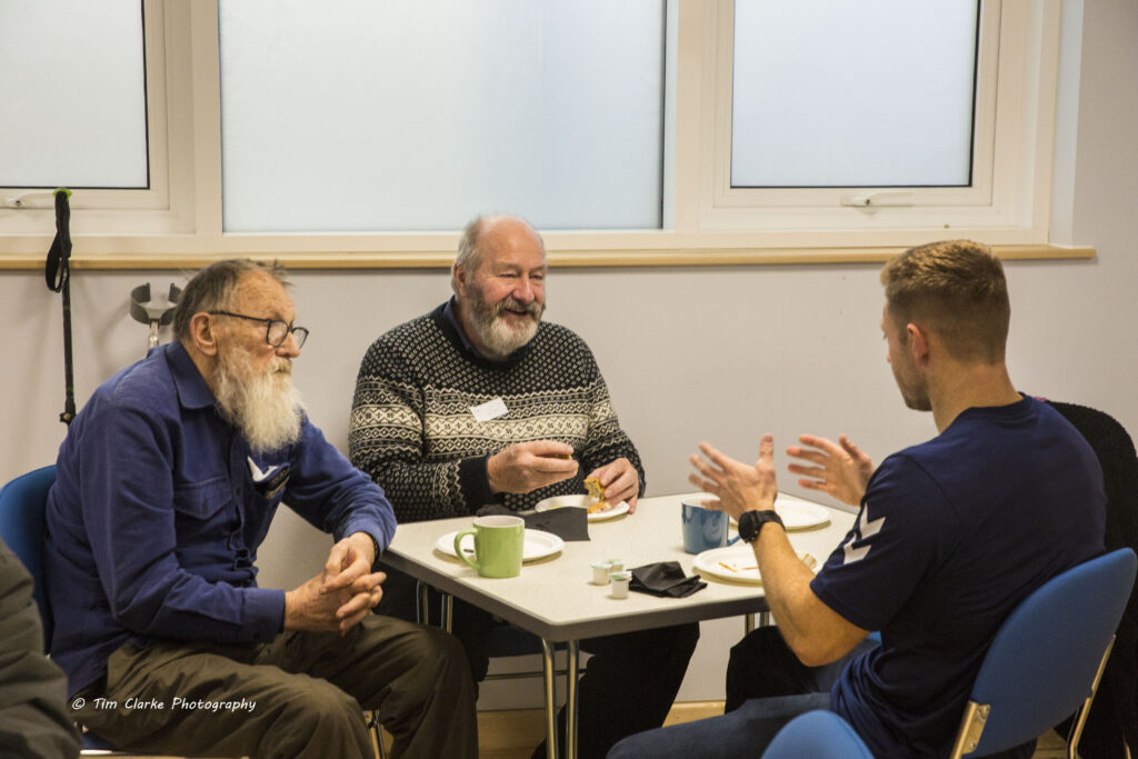 Two delegates (both white bearded gentlemen), chatting with Rob Oliver. All 3 are sat down at a table enjoying lunch.