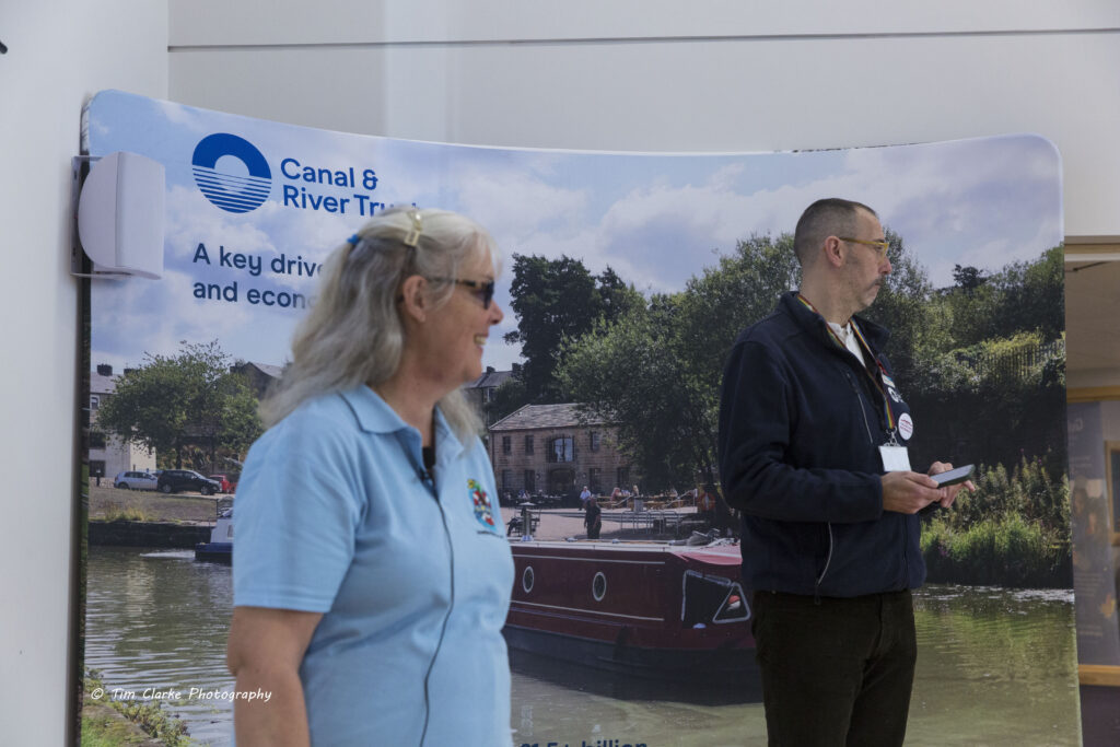 Tracey Clarke, wearing a light blue polo Shirt with the AWA logo on it to the left of Matthew Symmonds wearing dark blue fleece with the CRT logo. Behind is a large CRT banner with a canal scene.